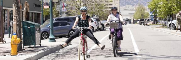 two women riding bikes downtown. One has her feet off the pedals and extended to the side in a playful manner. The other is following behind smiling.